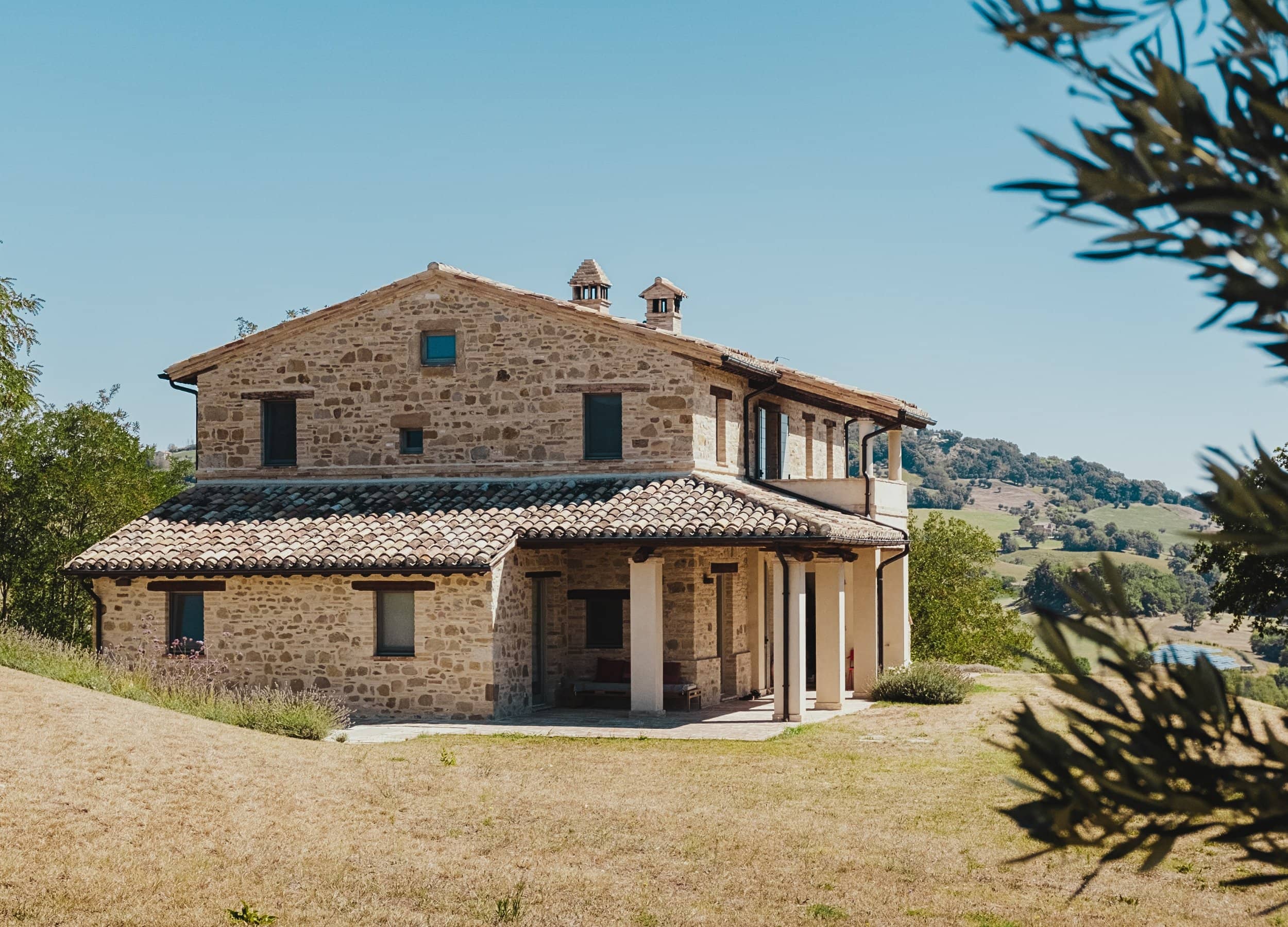 Stone house on a meadow landscape with covered entrance area. The sky is cloudless and in the distance are trees and a hilly terrain.