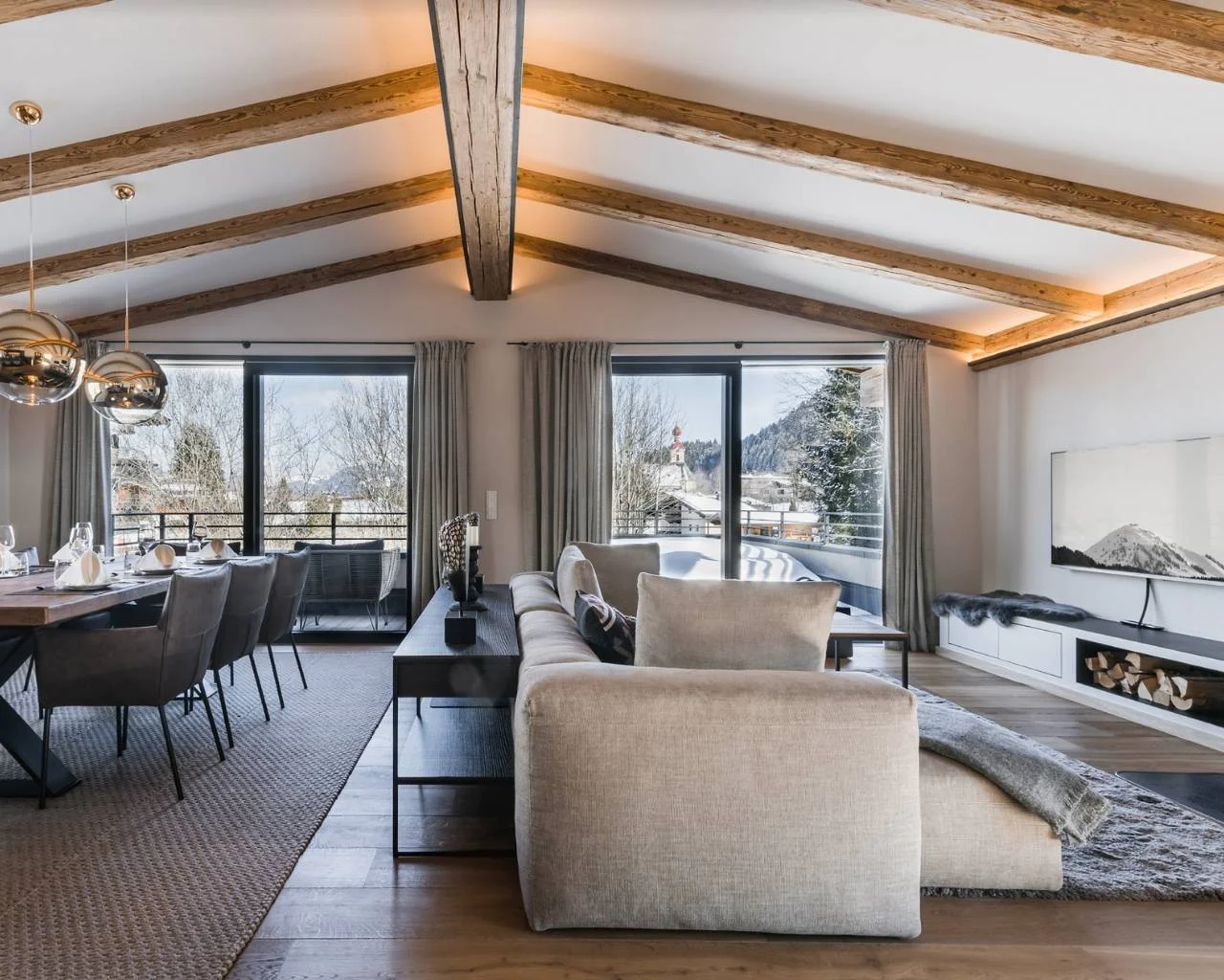 Elegantes Wohn-/Esszimmer mit direktem Zugang zum Balkon und Aussicht auf die Berge. Sichtbare, passive beleuchtete Dachbalken. Interior Design in Holz und Grautönen.