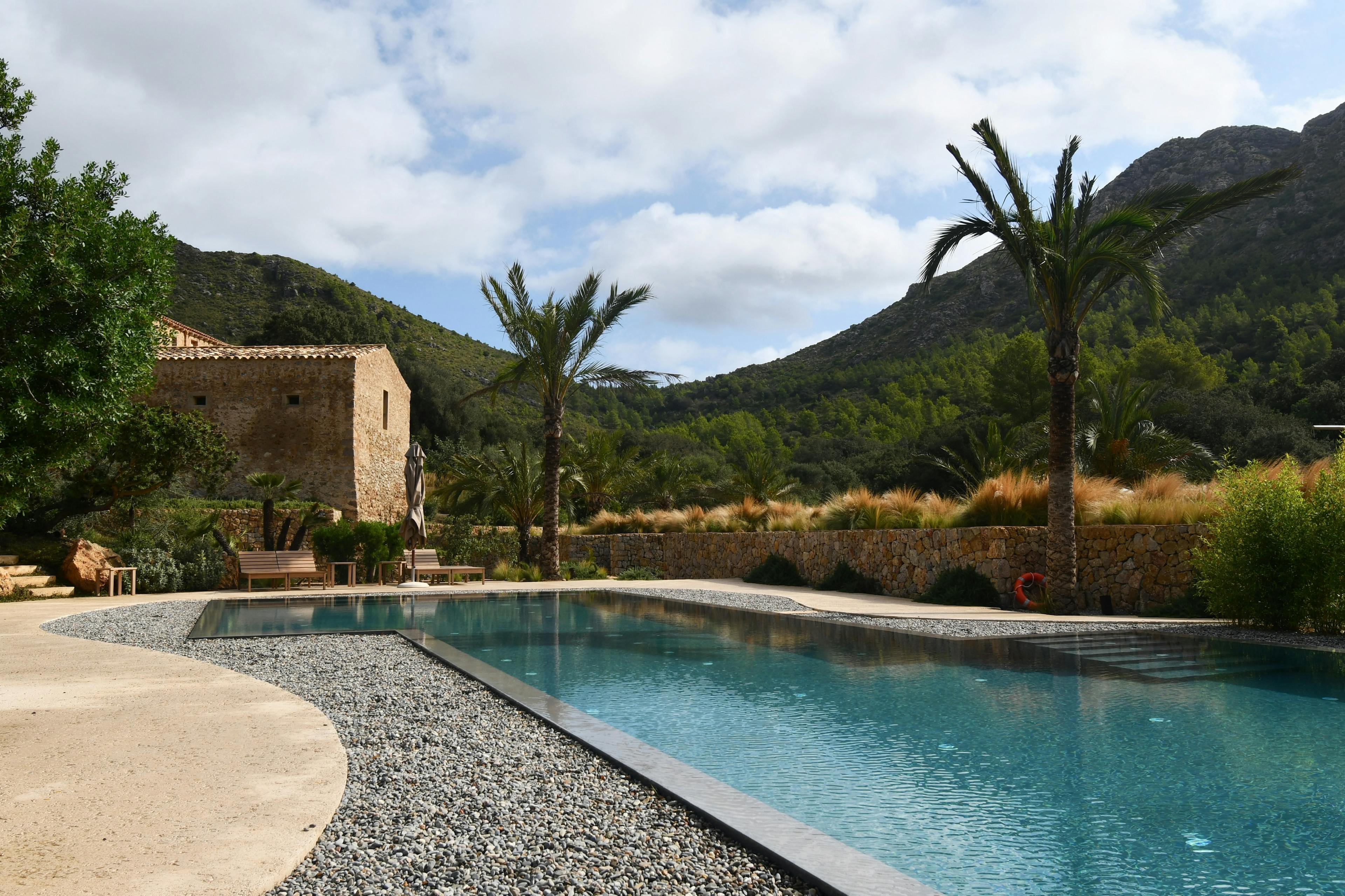 This mediterranean stone terrace is surrounded by the green of the mountains and a little stone wall. It has an embedded Pool and two palm trees.