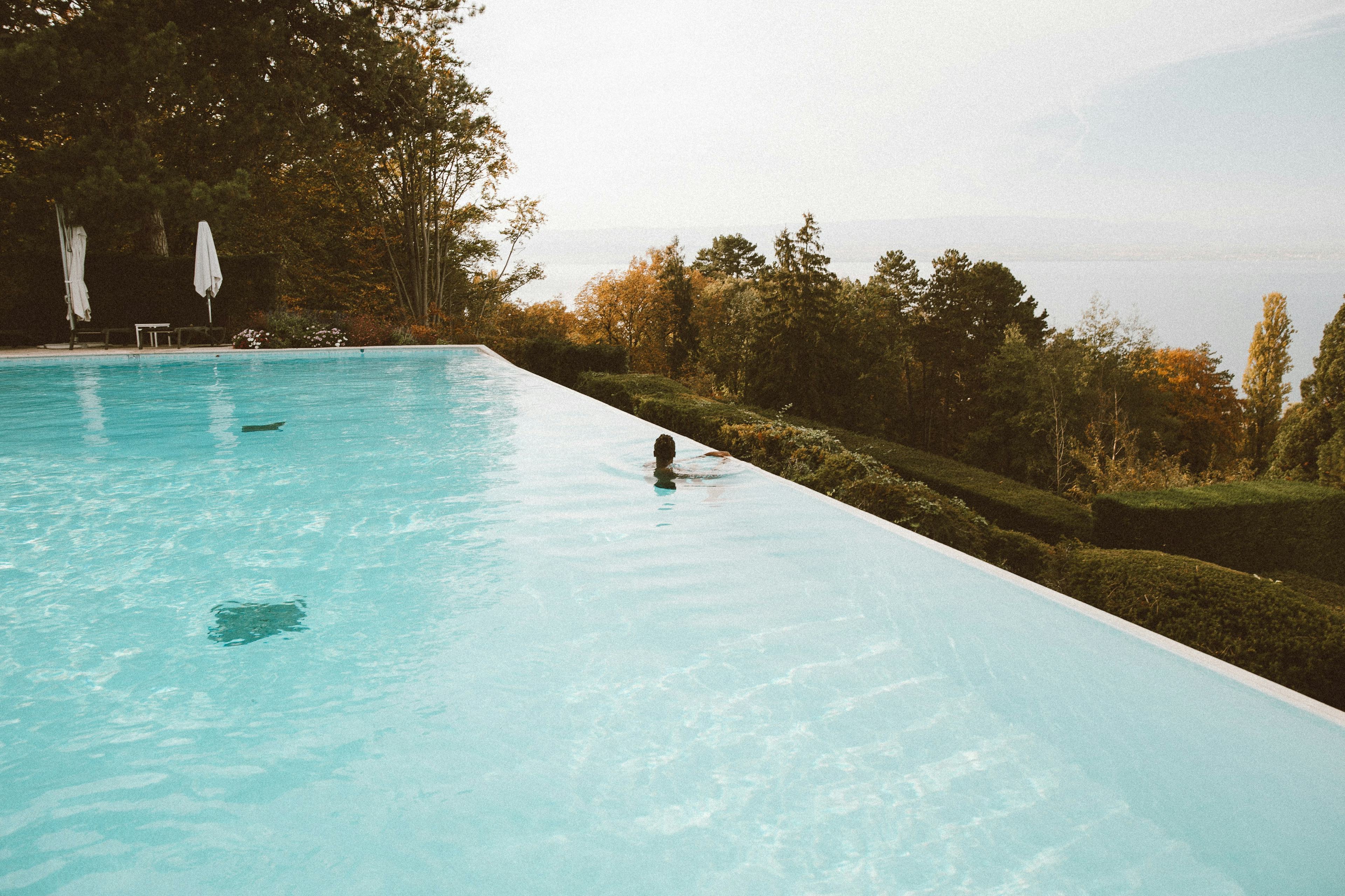 Large infinity pool with turquoise water in which a man on the edge contemplates the view of hedges and trees.  At the edge of the pool are bright folded umbrellas.