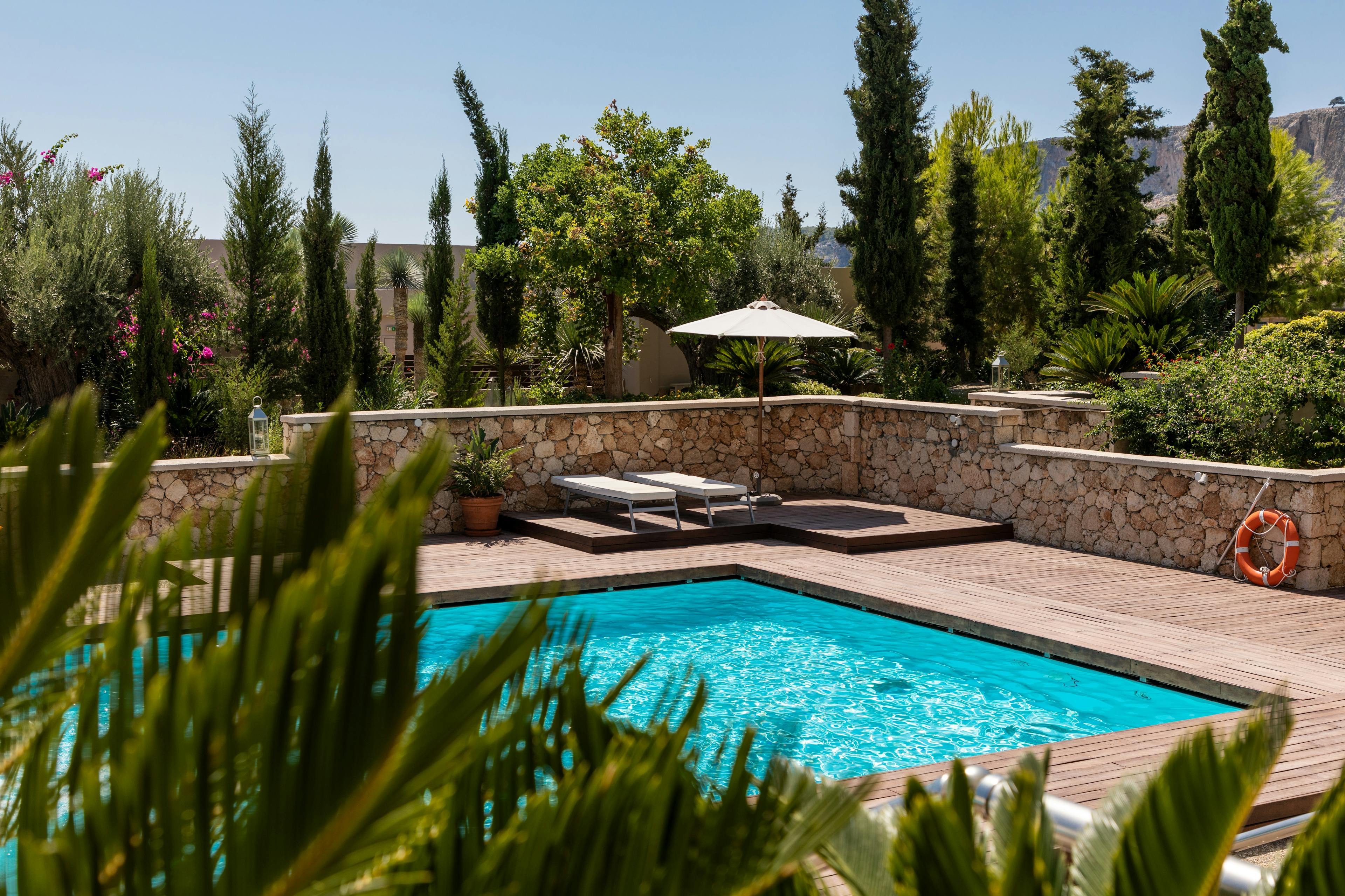 Privater Pool geschützt von einer mediterranen Steinmauer, eingelassen in die Hölzerne Terrasse. Leicht versteckt durch die umliegende Bepflanzung. Leichte Erhöhung in der Ecke mit 2 Sonnenliegen und einem Sonnenschirm.